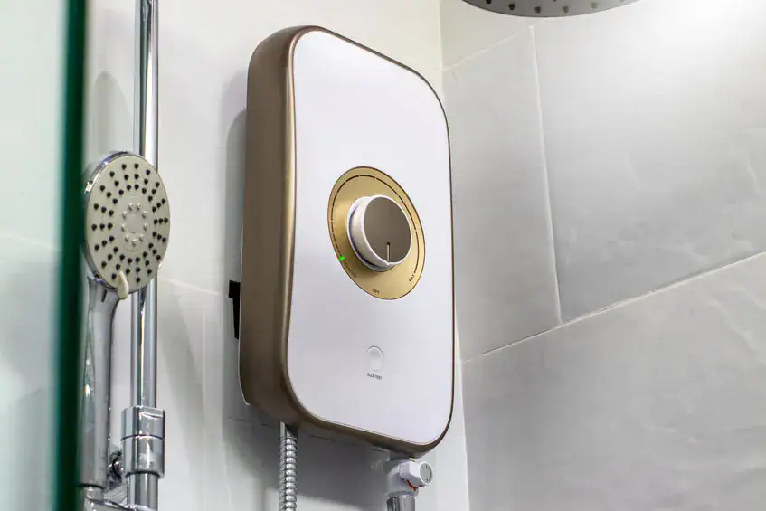 A modern electric shower water heater with a white and gold design is mounted on a tiled bathroom wall next to a chrome handheld showerhead.