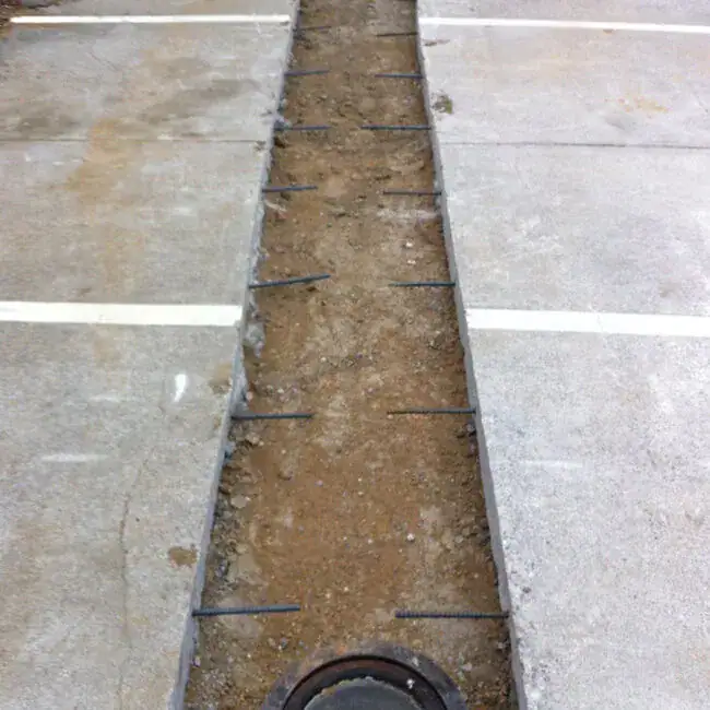 A parking lot with a long, narrow trench dug in the middle of the concrete, exposing dirt and rebar, and bordered by white parking lines.