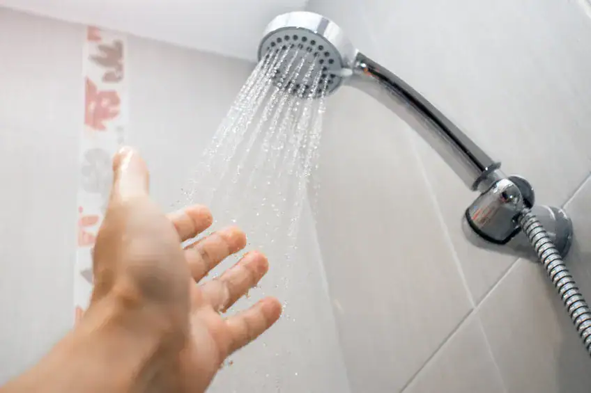 A person’s hand reaches toward running water from a wall-mounted showerhead in a tiled bathroom.