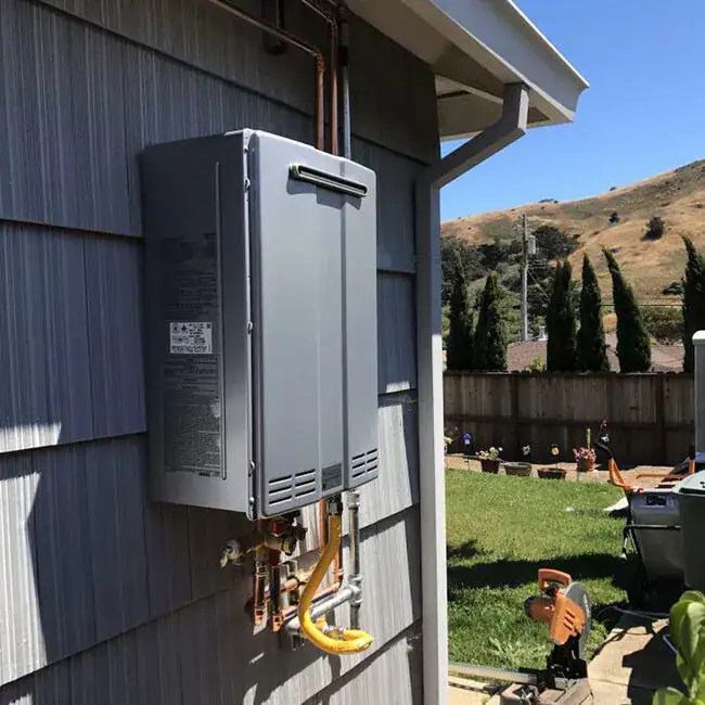 A tankless water heater is mounted on the exterior wall of a house in a backyard. A yellow hose and various pipes are connected to it. Trees, a lawn, and hills are visible in the background.