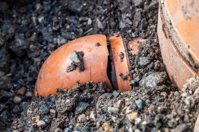 A broken orange clay pot partially buried in loose gravel and rocks, with the top half separated from the bottom half and resting at an angle.