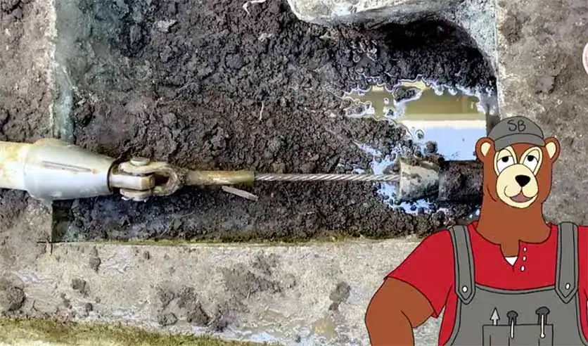 A pipe inspection tool is inserted underground, surrounded by wet soil, with a cartoon bear wearing overalls and a cap labeled "SB" standing on the right side of the image.