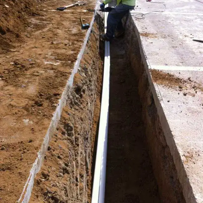 A worker installs a long white PVC pipe in a deep trench dug beside a concrete surface, with soil piled along the edge.