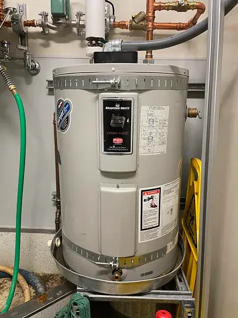 A water heater with various labels and instructions is installed in a utility area with visible copper piping, a green hose, and part of a yellow caution sign nearby.