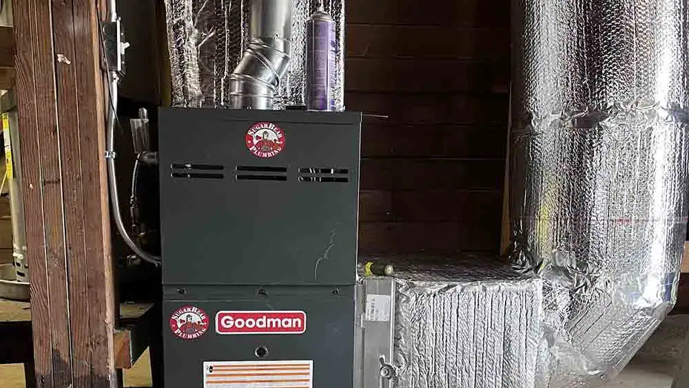A Goodman furnace unit installed indoors, surrounded by shiny, insulated ductwork and wooden beams. The furnace has manufacturer labels and warning stickers on its front panel.