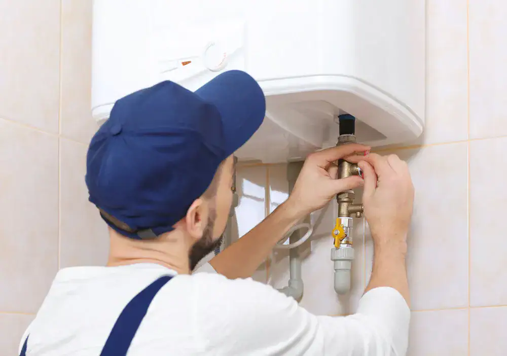 A plumber wearing a blue cap and white shirt adjusts valves on a white water heater mounted on a tiled wall.