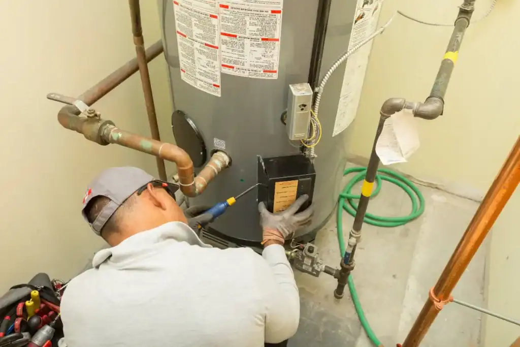 A technician wearing a gray cap, gloves, and a light gray shirt uses a tool to service a water heater surrounded by copper pipes in a utility room.