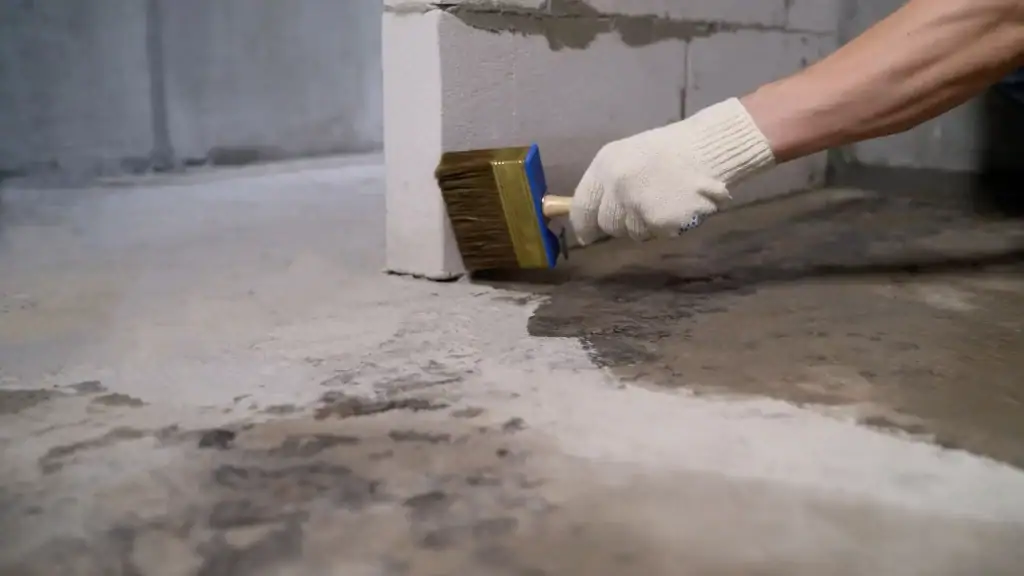 A person wearing a white glove uses a wide paintbrush to apply sealant or waterproofing treatment to the corner of a concrete floor and wall.