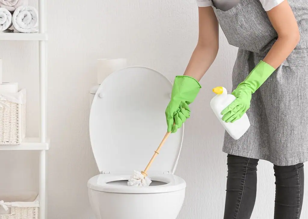 A person wearing green rubber gloves and a gray apron cleans a toilet with a brush in one hand and a bottle of cleaning solution in the other. Towels and toilet paper are visible on shelves nearby.