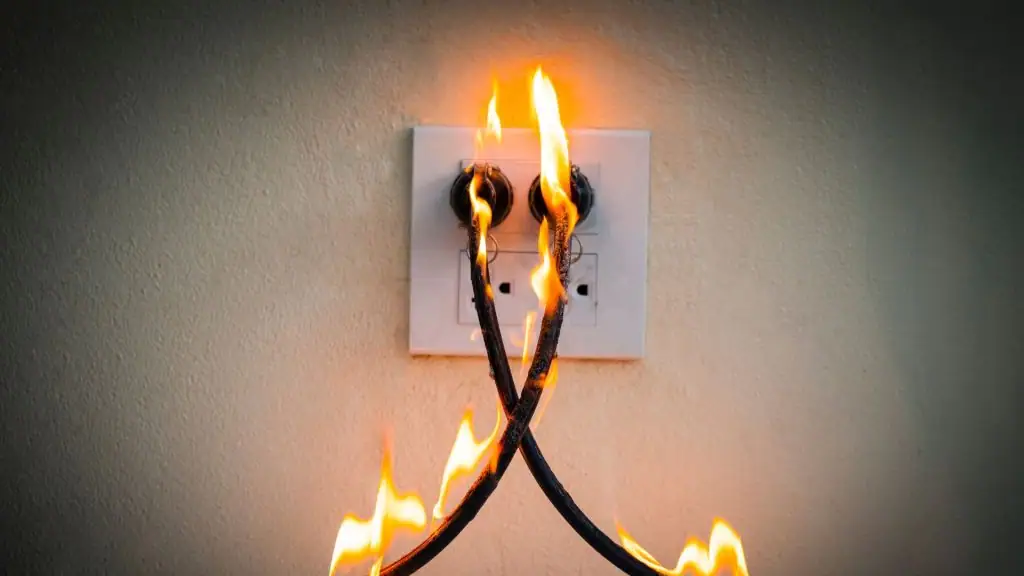 Two electrical cords plugged into a wall outlet are on fire, with visible flames along the wires and scorch marks on the outlet against a plain wall.