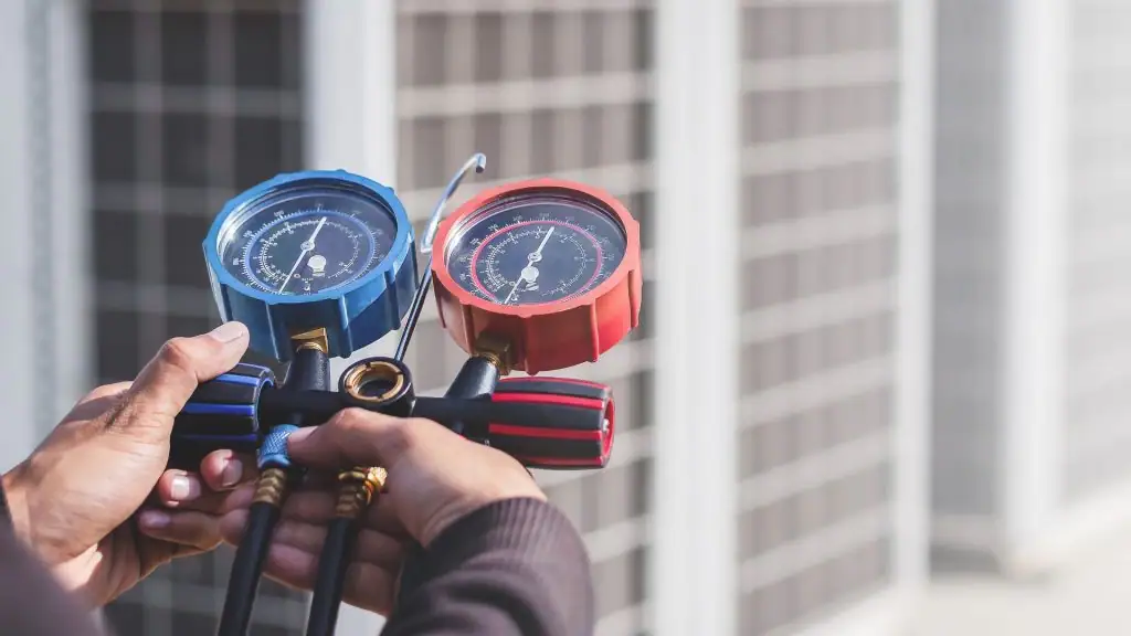 A person holds a manifold gauge set with blue and red dials, used for measuring pressure in air conditioning systems, with a blurred background of HVAC units.
