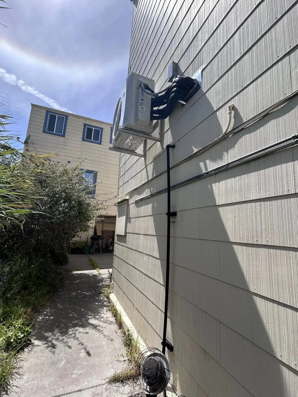 An outdoor air conditioning unit is mounted high on the side of a beige building, with black cables running down the wall. A small fan sits on the ground below, and a neighboring building is visible in the background.