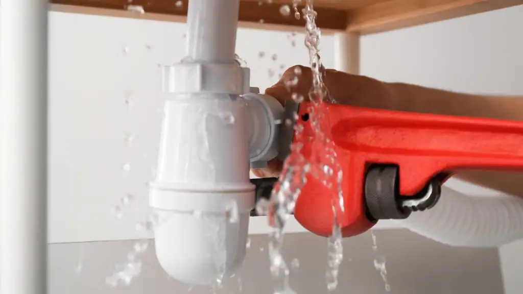 A person uses a red pipe wrench to tighten a leaking white pipe under a sink, with water spraying out around the connection.