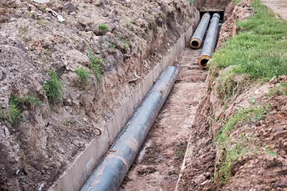 Large utility pipes are laid in a trench dug into the ground, surrounded by soil and patches of grass, likely as part of an underground infrastructure or construction project.