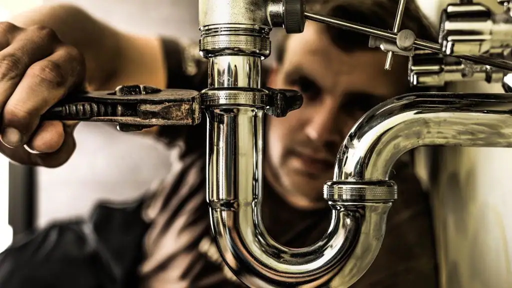 A plumber uses a wrench to tighten a shiny metal pipe under a sink, focusing intently on the plumbing work in a close-up view.