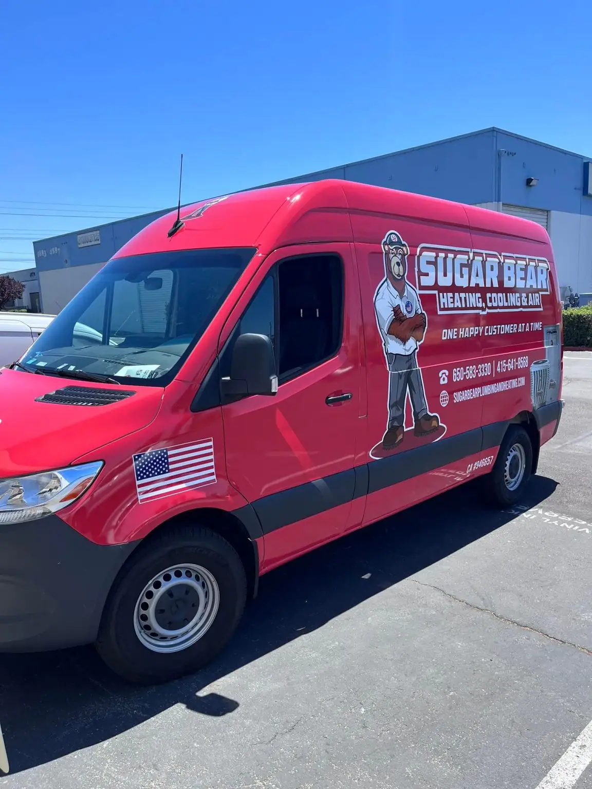 A red van for Sugar Bear Heating, Cooling & Air is parked outside a building. The van has a cartoon bear mascot, company name, phone numbers, and an American flag decal on the front door.