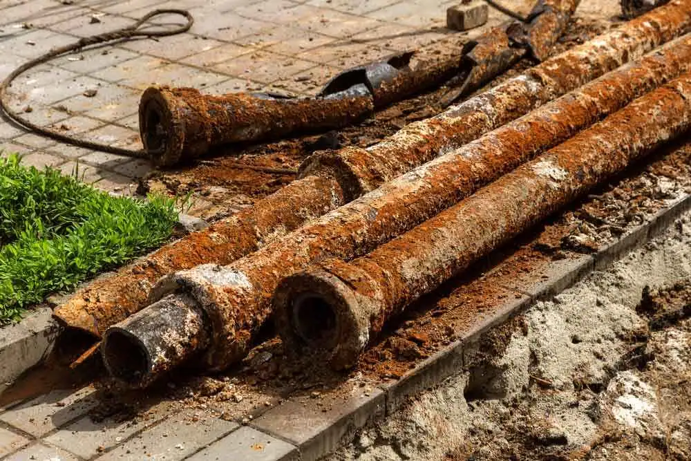 Several long, heavily rusted metal pipes lying on a sidewalk next to a patch of green grass and some broken pavement.