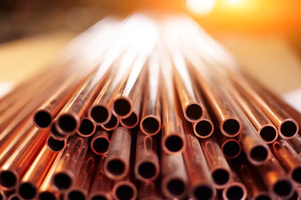 A close-up view of shiny, stacked copper pipes with a blurred, sunlit background. The pipes are arranged with their open ends facing the camera, showing their circular cross-sections.