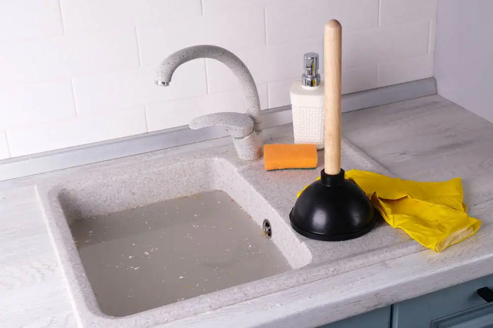 A kitchen sink filled with dirty water, a plunger, yellow rubber gloves, a sponge, and a soap dispenser are on the countertop nearby.