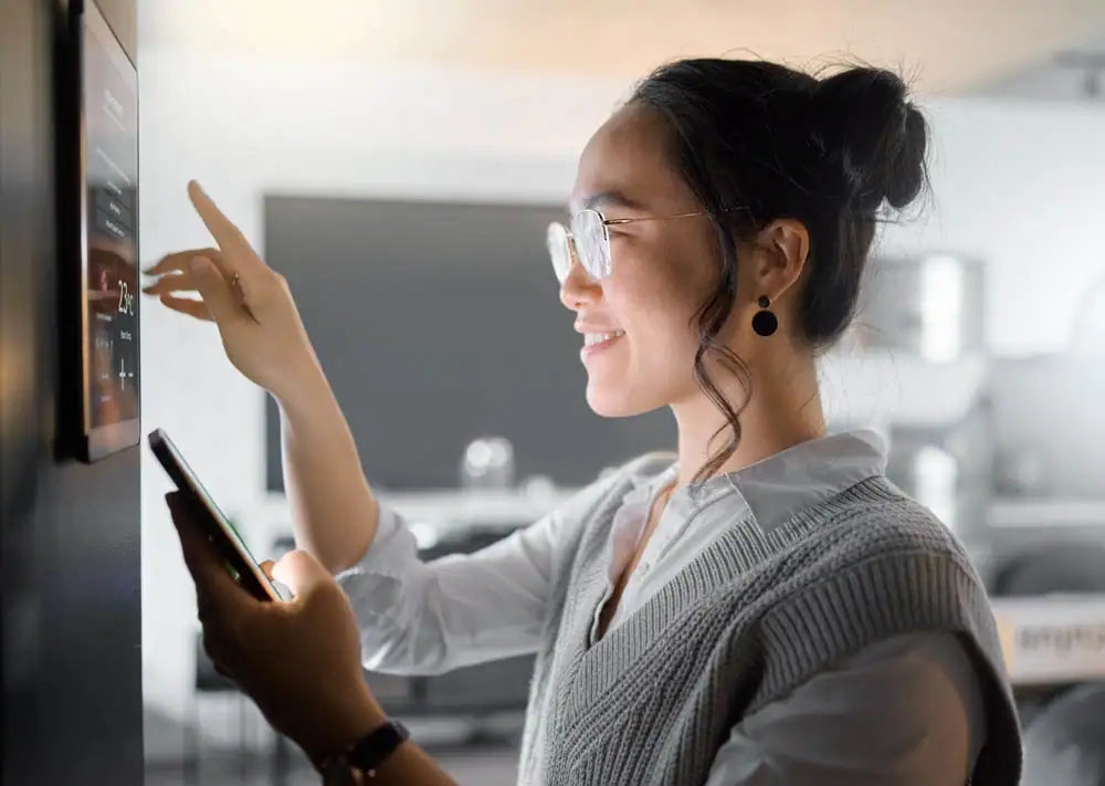 A woman wearing glasses smiles while adjusting a smart home control panel on the wall with one hand and holding a smartphone in the other. The room behind her is softly lit and modern.