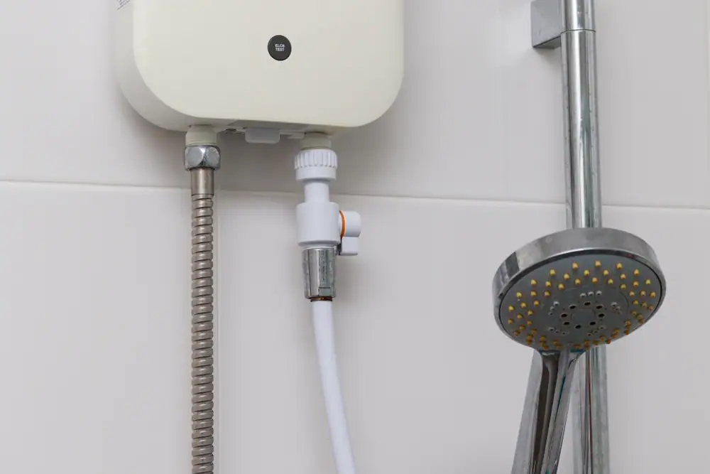Close-up of an electric water heater unit and a handheld showerhead mounted on a white tiled bathroom wall, with visible pipes and hose connections.